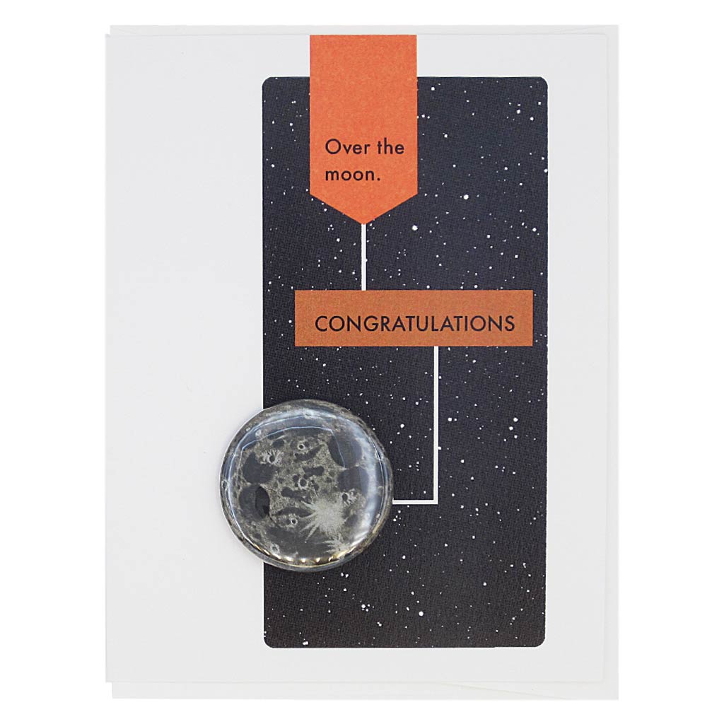 This congratulations card is designed to look like a page from a vintage science textbook with a black background of a starry sky. The text reads, ‘Over the Moon’ with an arrow to the text ‘Congratulations’ with an arrow to a 2¼” button of the Moon that can be taken off and worn by the recipient. Card measures 4¼” x 5½”, comes with a white envelope & is blank inside. Designed by The Regional Assembly of Text.